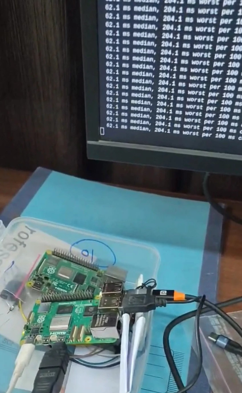 Two Raspberry Pi boards in a clear box on the bench, cabled up, with a monitor behind them printing the model's per-chunk timing during a live run.